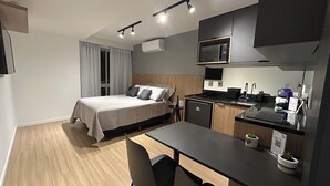 Una habitación de hotel compacta con cama, kitchenette, pequeña mesa de comedor y una silla.