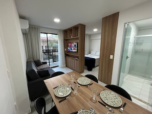 Un comedor con una mesa de madera dispuesta para cuatro, un televisor al fondo y un box de ducha con paredes de vidrio.