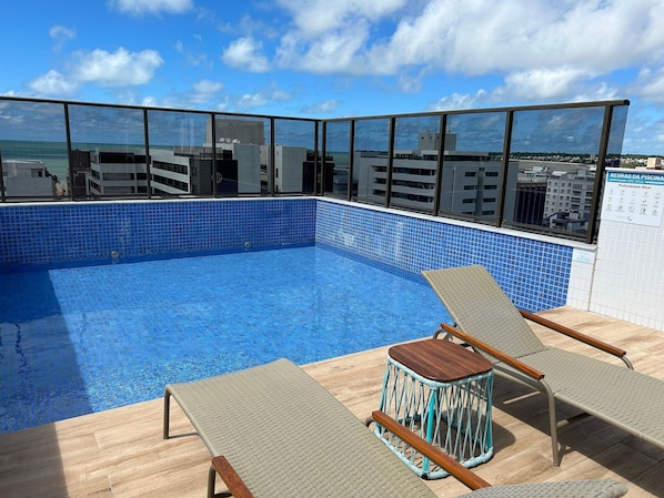 A rooftop swimming pool with clear blue tiles, two lounge chairs, and a view of the cityscape.