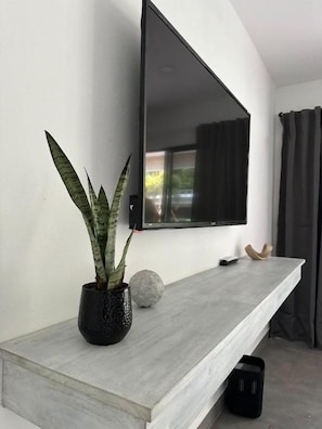 A modern living room with a flat-screen TV mounted on a wall, a grey console table, a potted plant, and a stone sculpture. - Hotel Good Life (Coco)