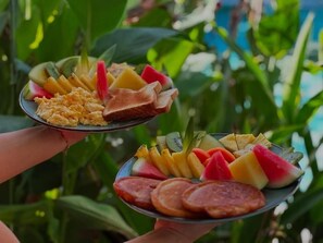 Two plates of food with fruits and scrambled eggs, held by a person. - Hotel Good Life (Coco)