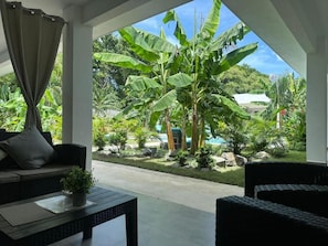 An outdoor seating area with wicker furniture, a potted plant, and a view of a lush garden with banana trees and a swimming pool. - Hotel Good Life (Coco)