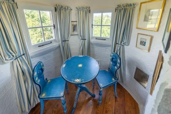 Una habitación con una mesa redonda azul y cuatro sillas a juego, ventanas amplias con cortinas y cuadros en la pared.