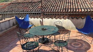 En terrasse med et rundt bord og stole, en stor parasol samt udsigt til et tag med fliser og grønt i baggrunden.