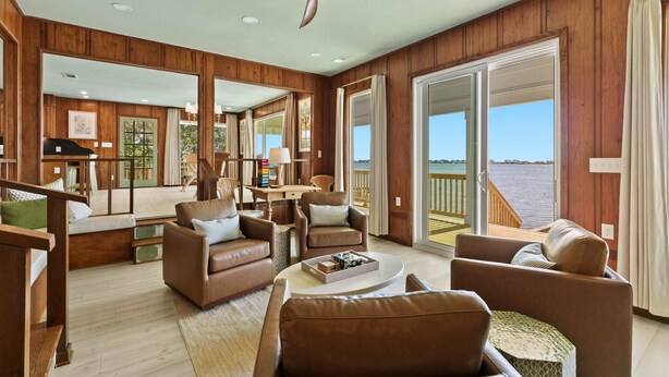A spacious living room with wooden paneling, leather furniture, a ceiling fan, and large sliding glass doors leading to a balcony with a view of the water.