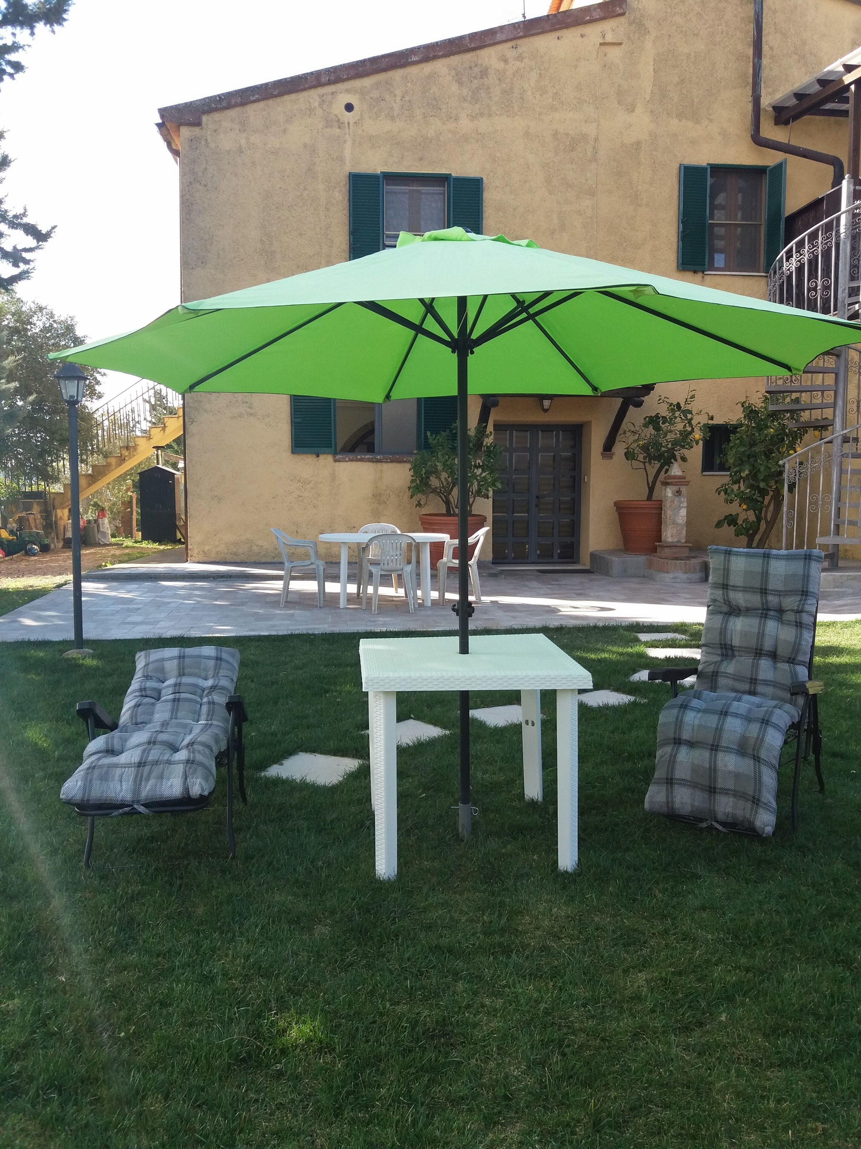 An outdoor seating area with a table and chairs under a green umbrella, a small lawn, and a building with a yellow facade and green shutters in the background.