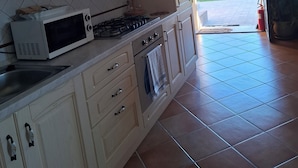 A kitchen with white cabinets, a microwave, stove, and oven. There is a view of a green landscape through an open door.