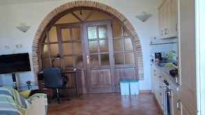 A room with a bed, desk, chair, and a kitchen area with cabinets and a stove.