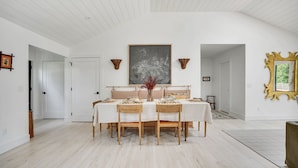 Un comedor con una mesa dispuesta para cuatro personas, rodeada de sillas, con un cuadro abstracto grande encima de la mesa y un espejo decorativo en la pared.
