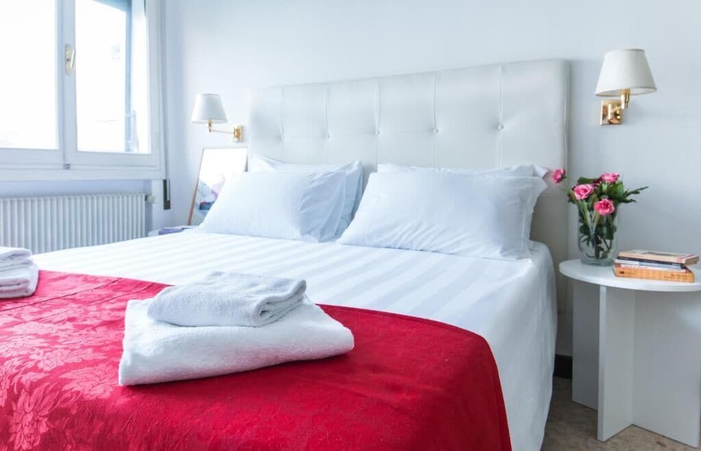 Quarto de hotel com uma cama grande, roupa de cama branca, colcha vermelha decorativa, mesa de cabeceira pequena com abajur e um vaso com flores.