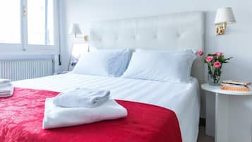 Quarto de hotel com uma cama grande, roupa de cama branca, colcha vermelha decorativa, mesa de cabeceira pequena com abajur e um vaso com flores.