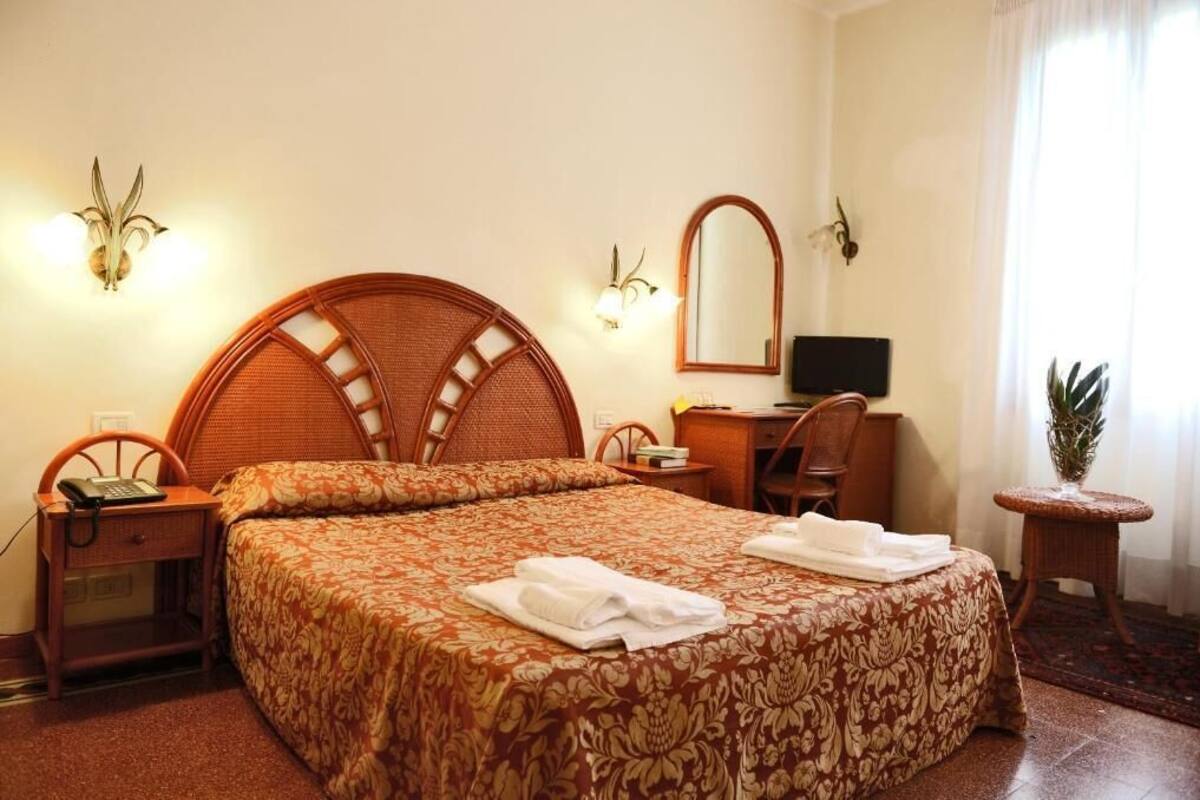 A hotel room with a large bed, a desk, a chair, a small table, and a potted plant.