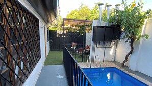 Une piscine entourée d’une clôture en métal noir, un petit jardin agrémenté d’une pergola en bois et un portail en métal noir.