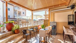 An outdoor seating area with tables and chairs, potted plants, and a bar area with stools. - Elena Hotel Apartments (Parga)