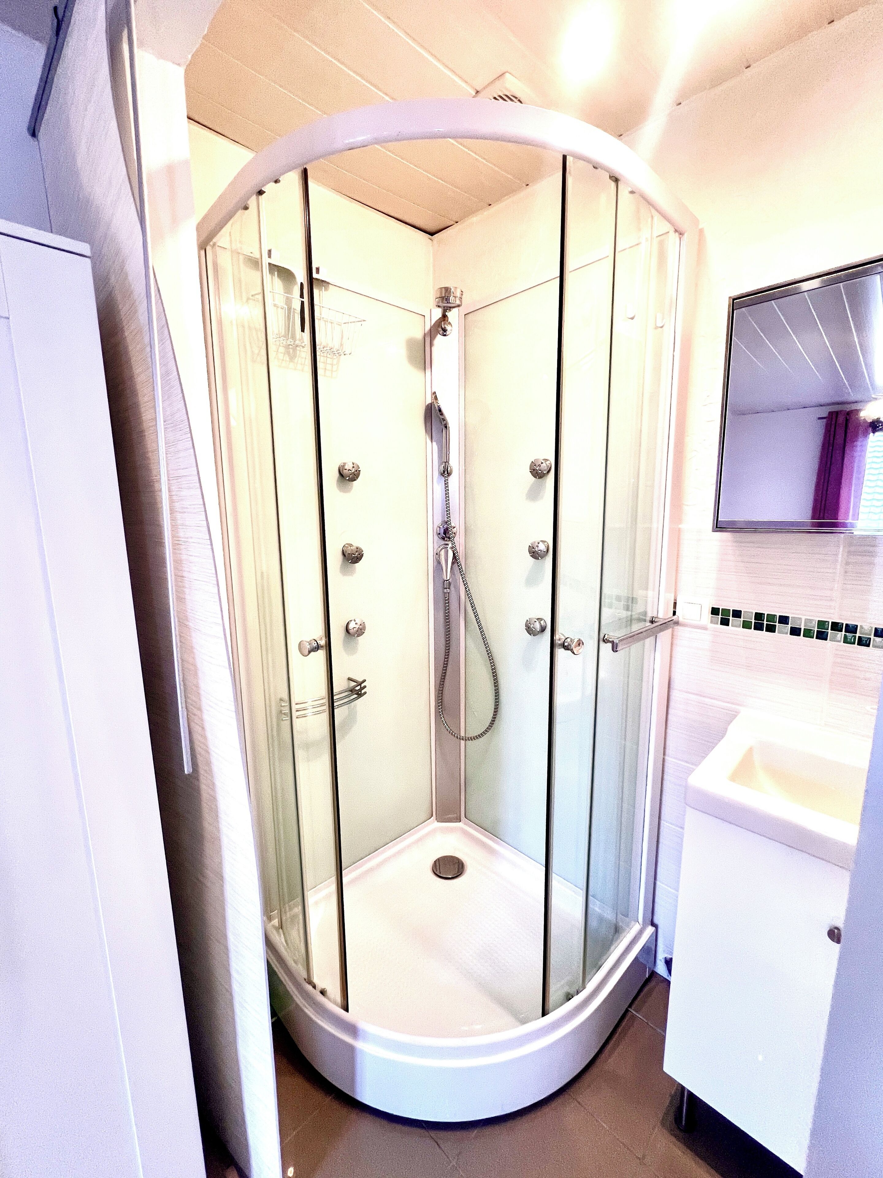 Une cabine de douche fermée par des parois en verre, dotée d’un pommeau de douche à main, d’un petit drain et d’un lavabo blanc avec un miroir au-dessus.