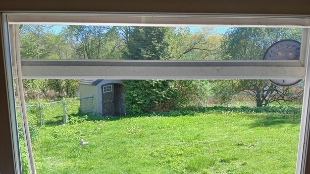 Una ventana con vista a un jardín cubierto de césped, un objeto de madera en el alféizar y un medidor de agua en la pared.