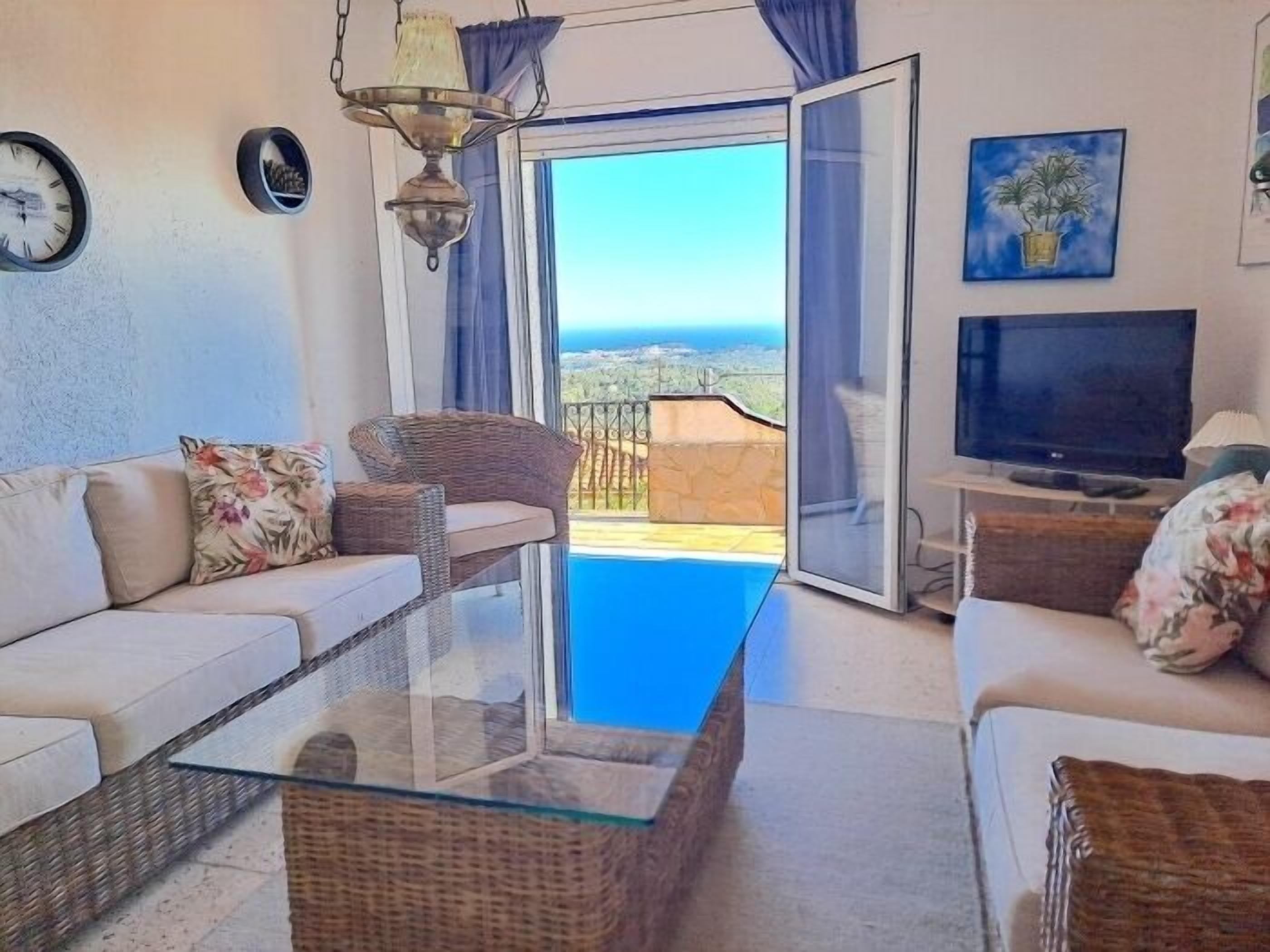 Un salon avec une table basse en verre, des meubles en osier, un téléviseur et une vue sur la mer.