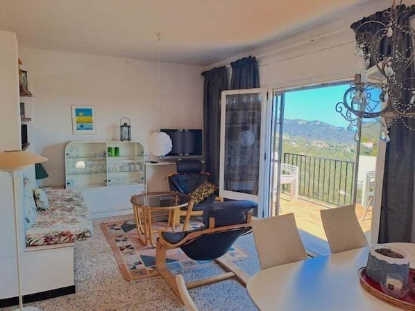 A living room with a sofa, a dining table, chairs, a TV, and a balcony with a view.