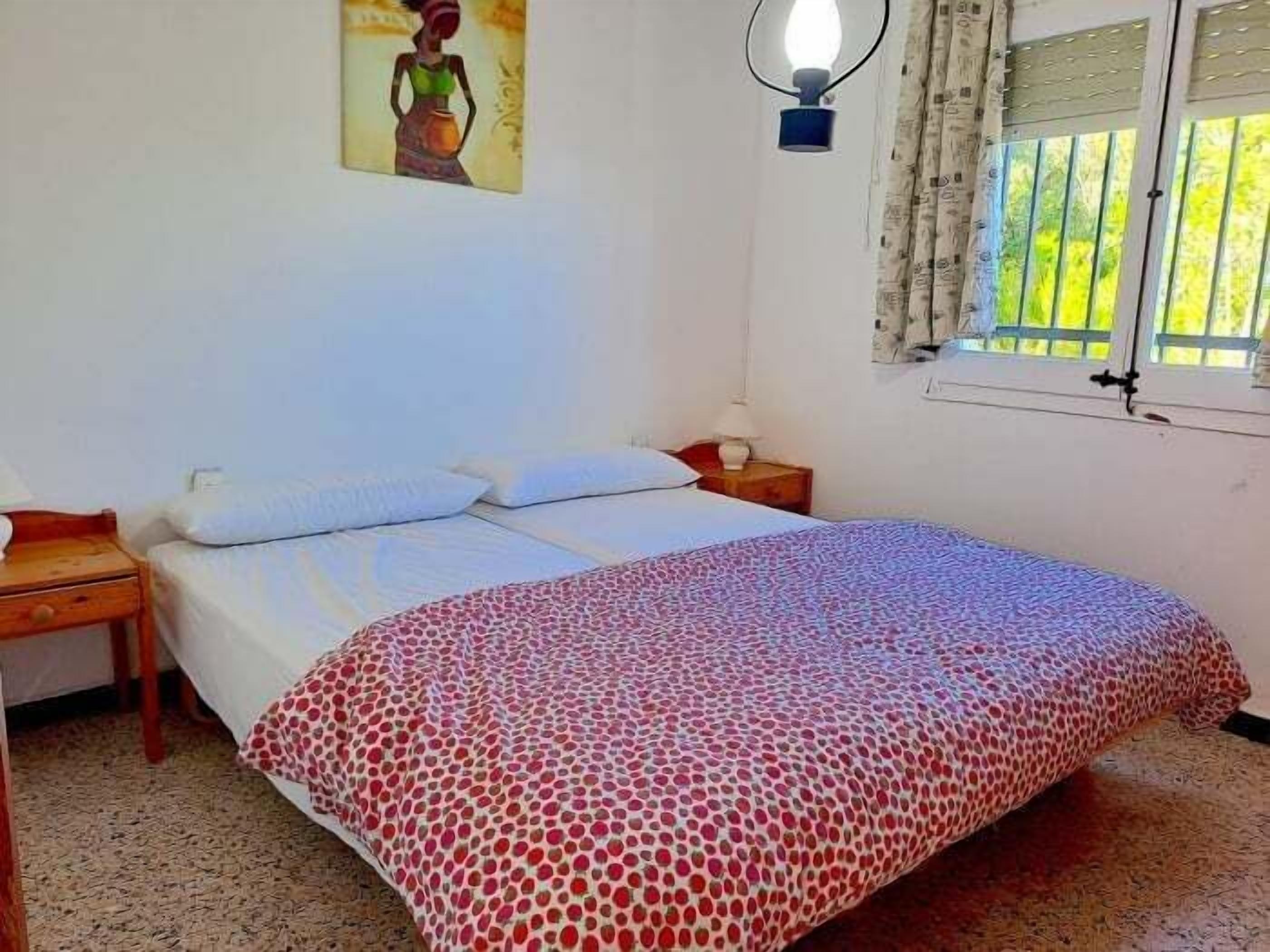 Une chambre à coucher comprenant un lit, une table de chevet, une lampe fixée au mur, une fenêtre avec des rideaux et un tableau accroché au mur.