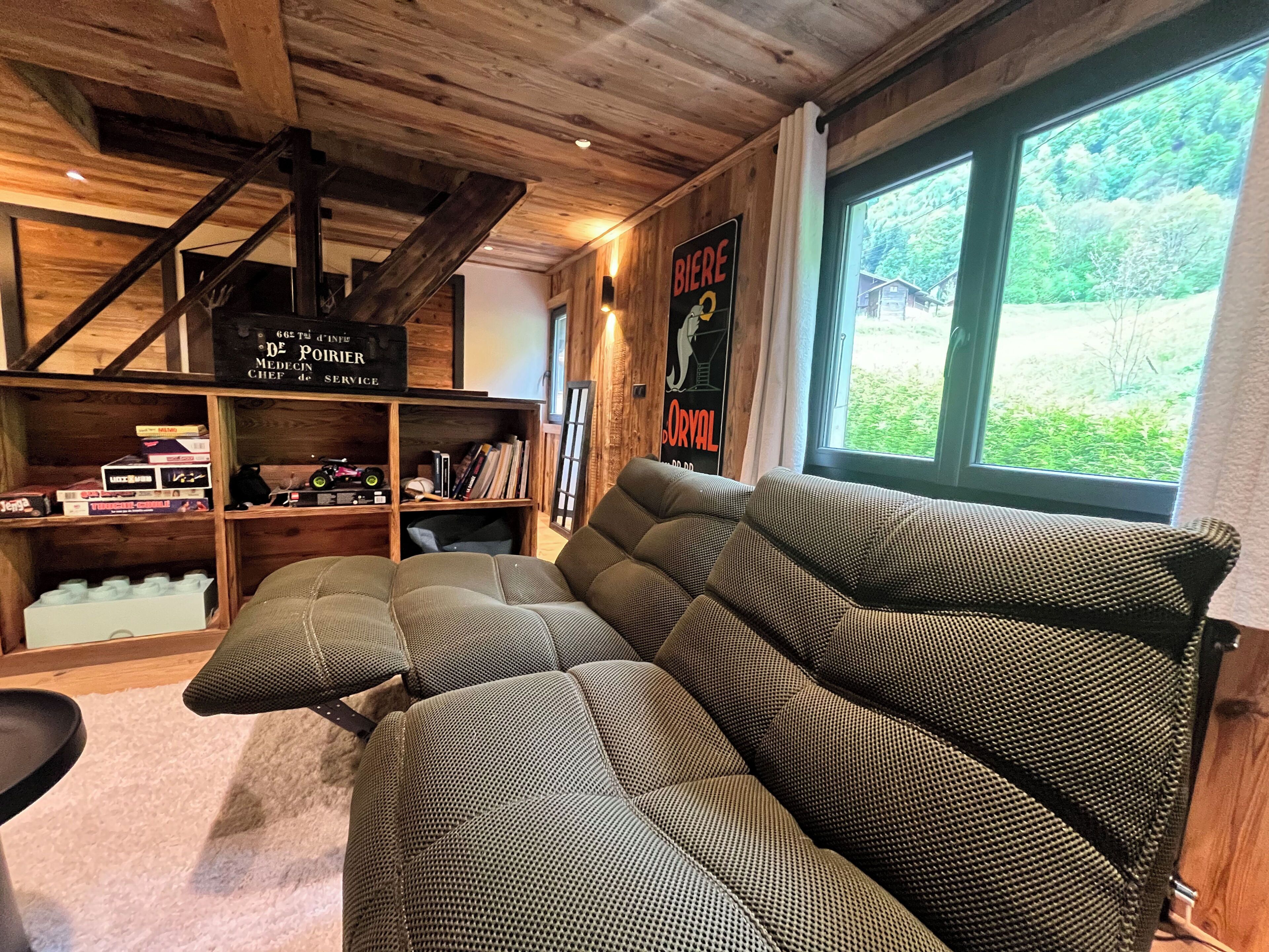 A cozy living room with a large sectional sofa, wooden beams on the ceiling, a bookshelf with various items, and a window with a view of greenery.