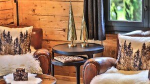 L’intérieur chaleureux d’un chalet avec des murs en bois, une table ronde sur laquelle est posé un jeu d’échecs, et deux chaises agrémentées de coussins aux motifs forestiers.