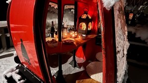 Un véhicule rouge, fermé, entièrement recouvert de neige, avec une table et des chaises à l’intérieur, éclairé par une lumière chaleureuse.