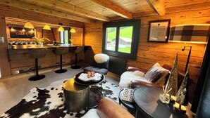 Un intérieur de chalet chaleureux avec un plafond en bois, un coin bar, un espace salon agrémenté d’un tapis en peau de vache et une fenêtre donnant sur un paysage verdoyant.