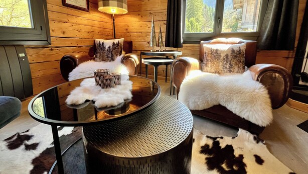 L’intérieur chaleureux d’un chalet avec des murs en bois, une table ronde recouverte d’un plaid en fourrure, un canapé agrémenté d’une couverture en fourrure et une fenêtre offrant une vue sur les arbres et les montagnes.