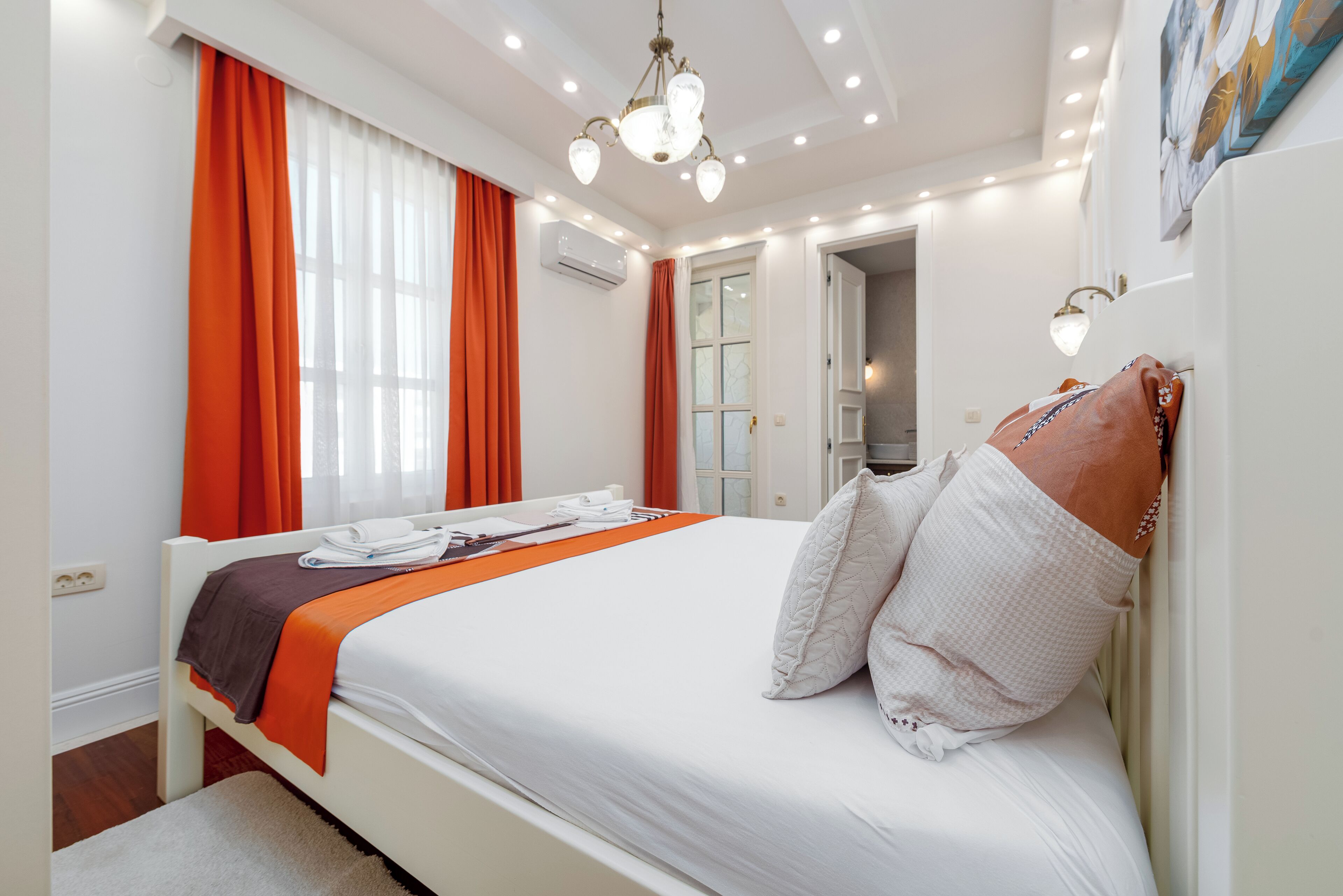 A hotel room with a neatly made bed, a large window with orange curtains, an air conditioning unit, and a hallway leading to another room.