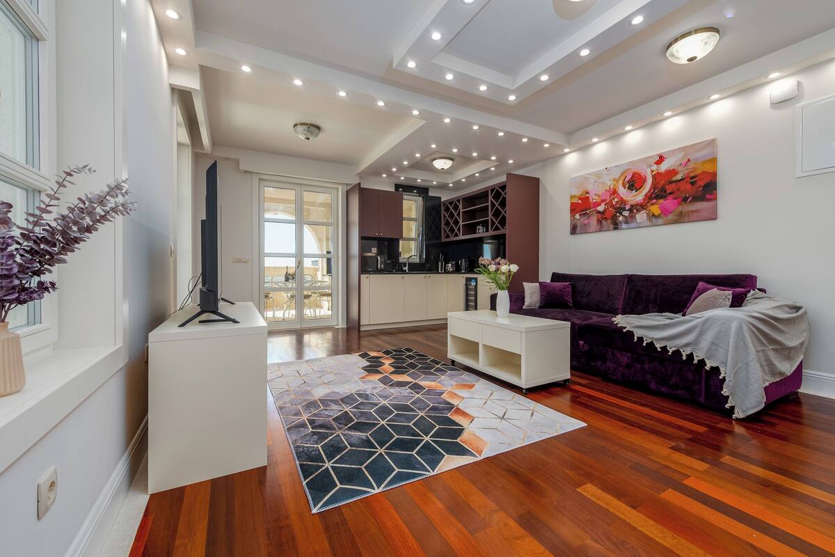 A modern living room with a wooden floor, white furniture, a purple sofa, and a colorful abstract painting on the wall.