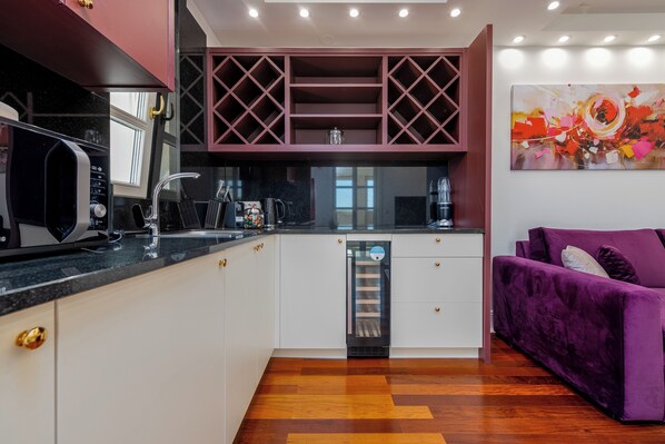 A compact kitchen with a dishwasher, a microwave, and a coffee maker. There is a purple sofa and a painting on the wall.