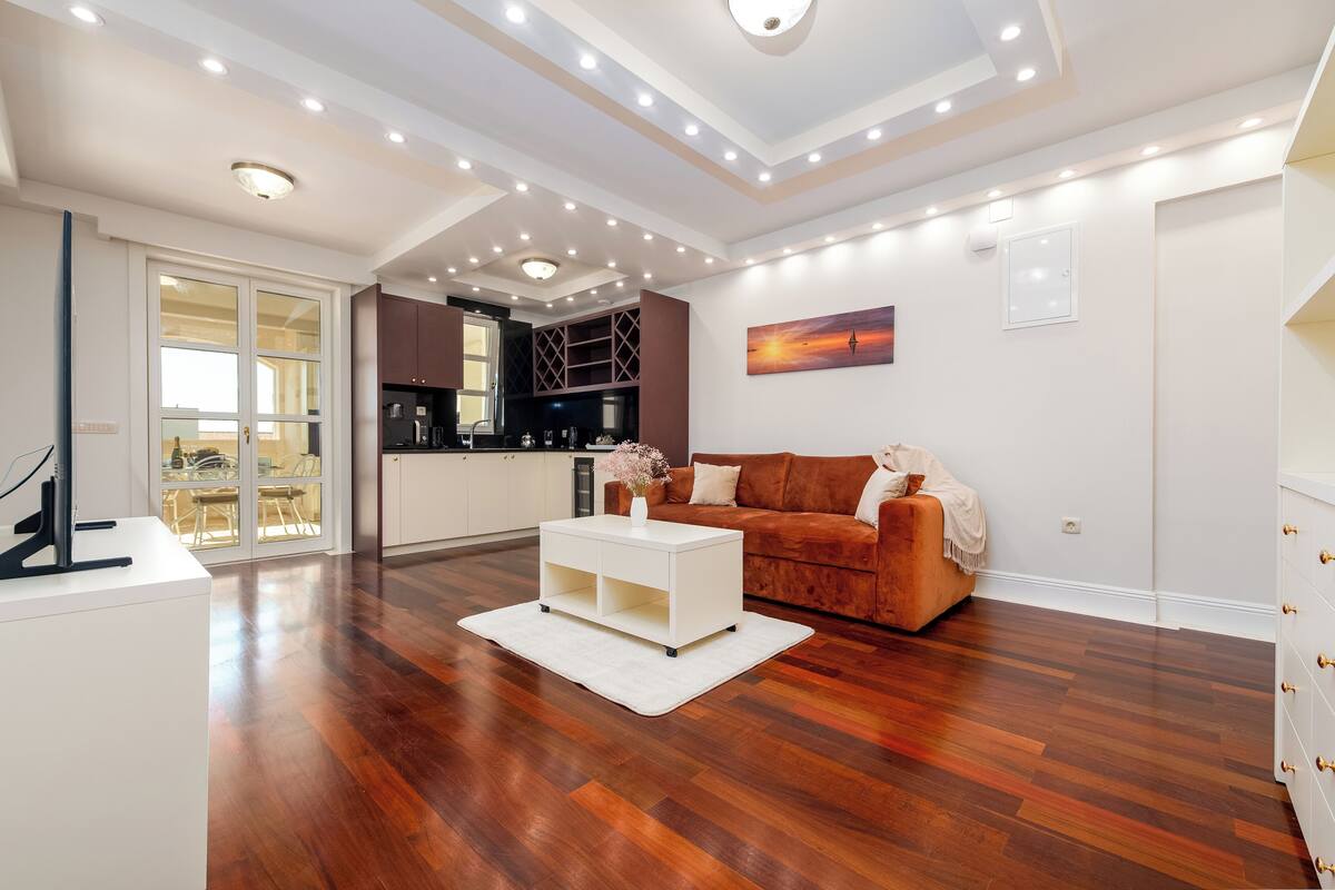 A spacious living area with a wooden floor, a white coffee table, a brown sofa, and a kitchen in the background.