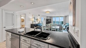 A modern kitchen with a black countertop, white cabinets, and a double sink. There is a dining area with a table and chairs visible through the kitchen.