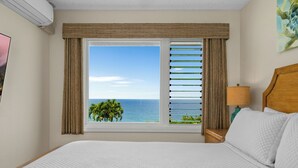 Een hotelkamer met een groot bed, een nachtkastje met lamp, een raam met zonwering en uitzicht op de oceaan en palmbomen.
