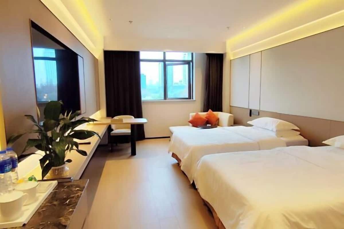 A hotel room with two beds, a desk, a chair, a potted plant, and a window with curtains.