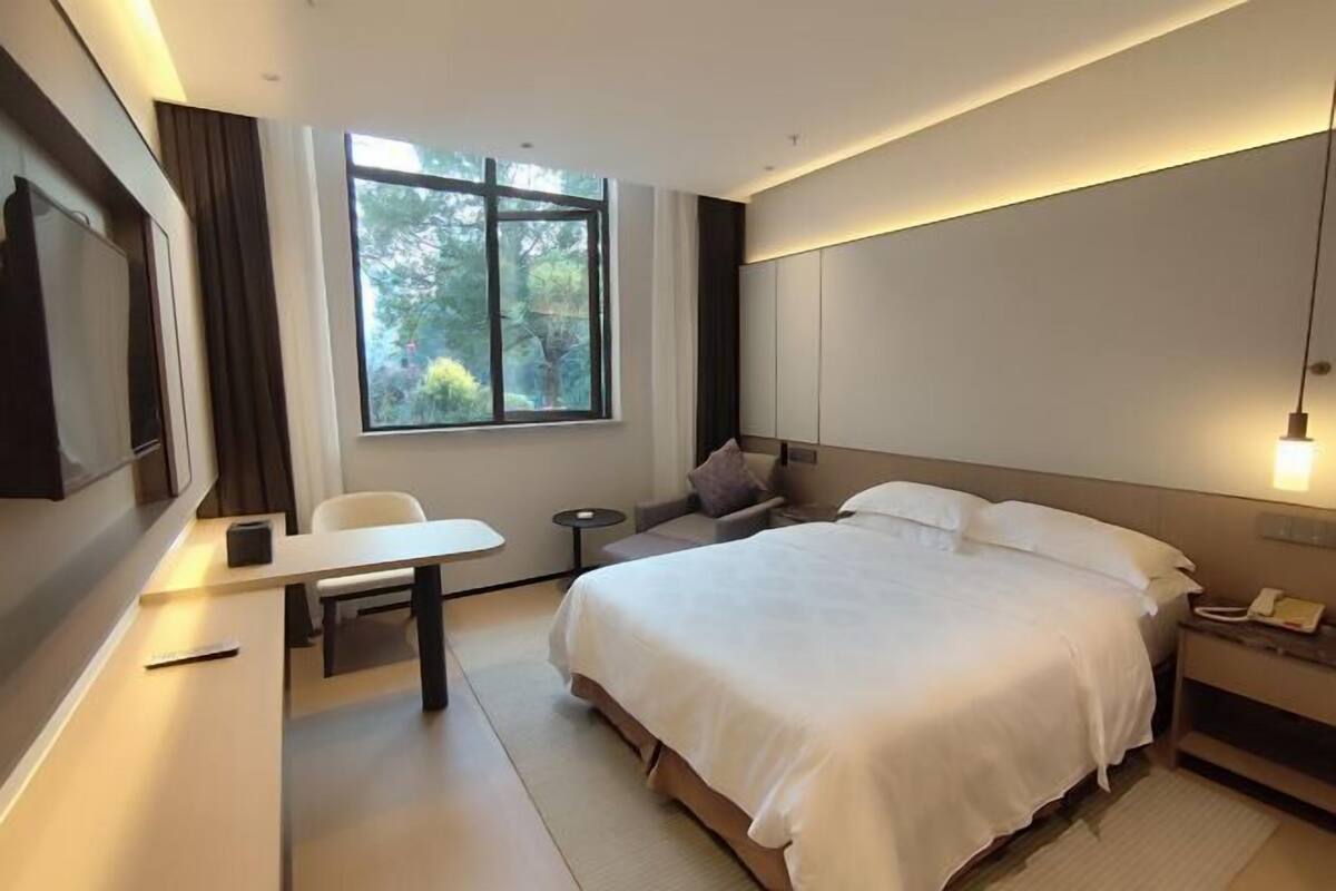 A modern hotel room with a large bed, a desk, a chair, a small table, a nightstand, and a window with a view of trees.