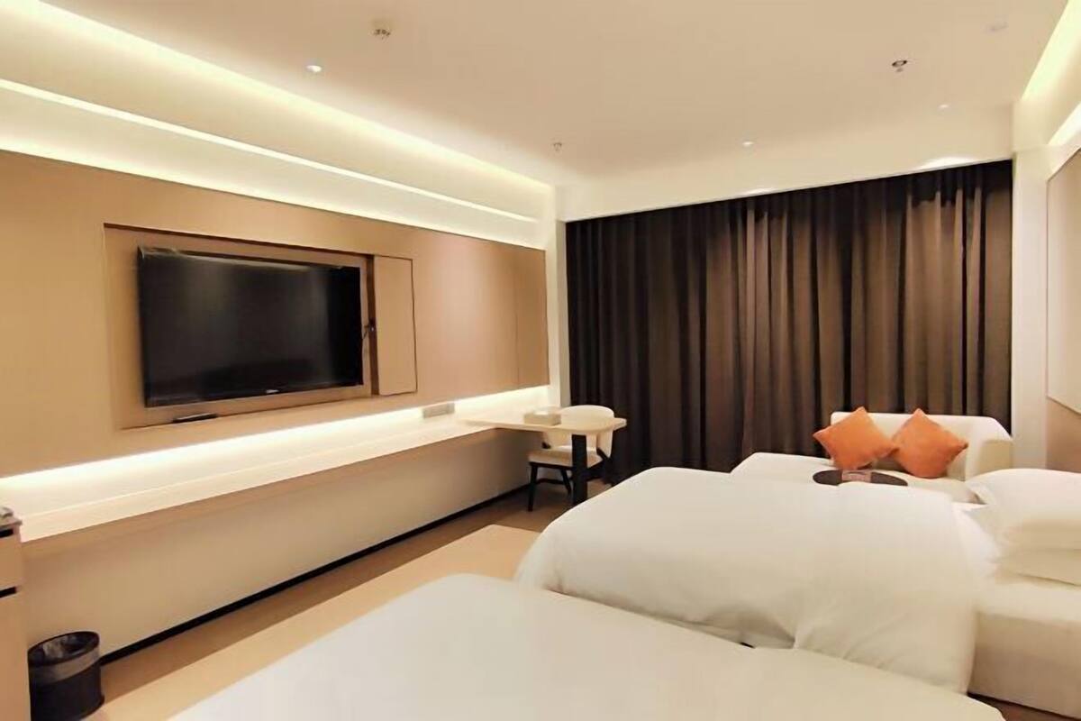 A modern hotel room with a flat-screen TV mounted on the wall, a small table with a chair, and a large bed with white bedding and orange pillows.