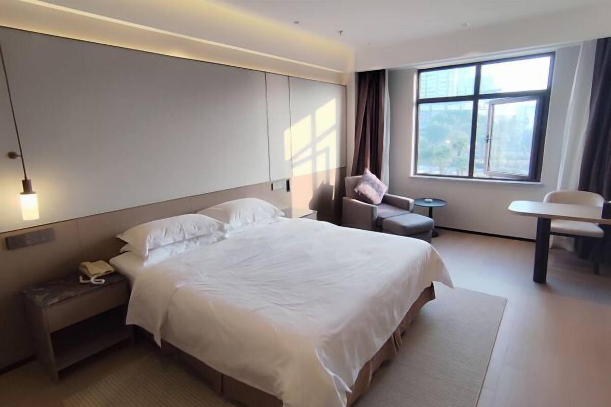 A modern hotel room with a large bed, a small sofa, a desk, and a window with a city view.