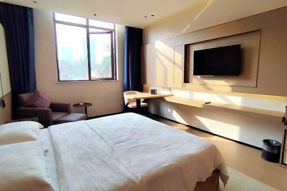 A hotel room with a large bed, a desk, a chair, a TV mounted on the wall, and a window with curtains.