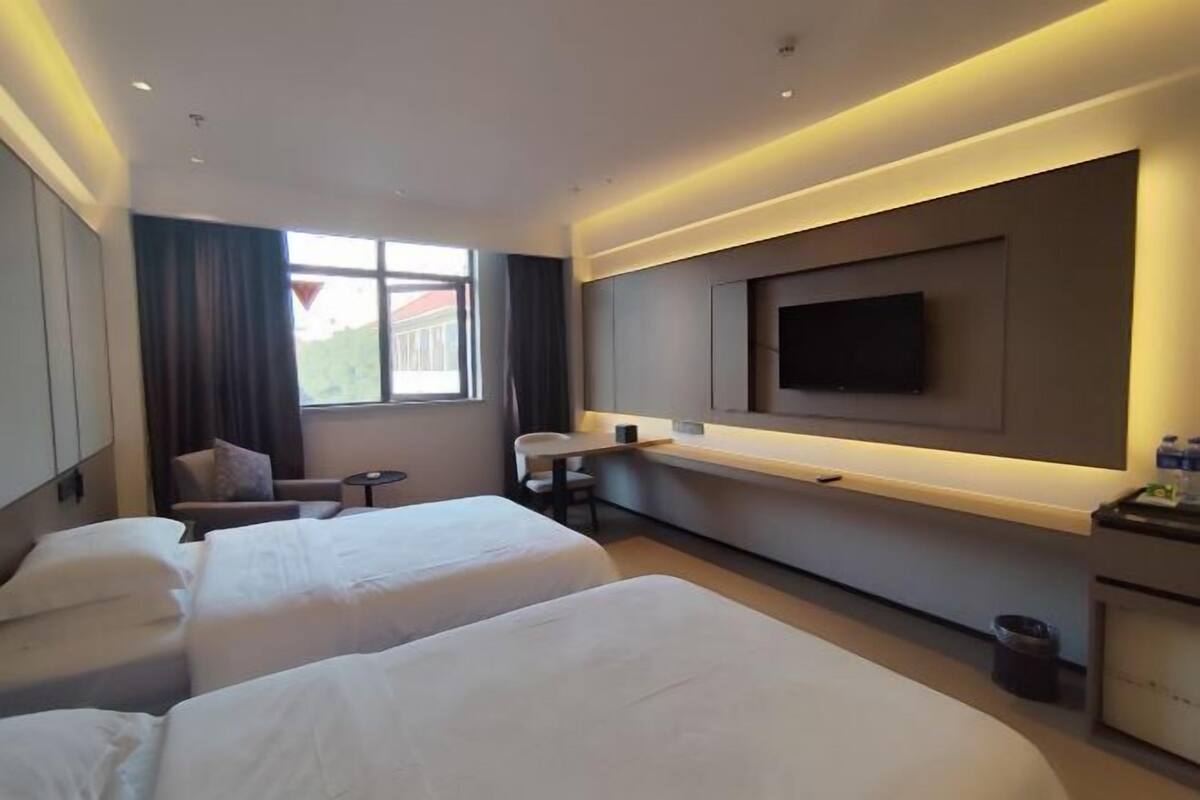 A hotel room with two beds, a desk, a TV mounted on the wall, and a window with curtains.