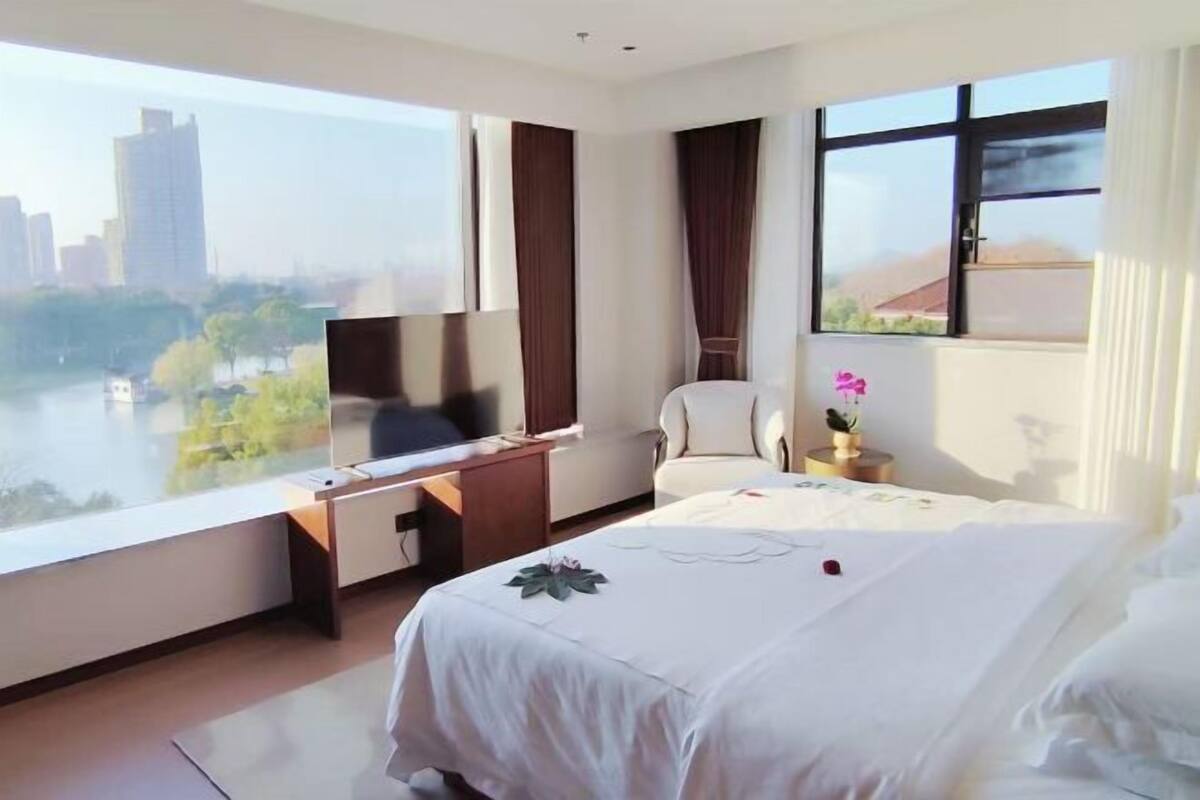 A hotel room with a bed, a desk, a chair, a TV, and large windows with a view of a cityscape and river.