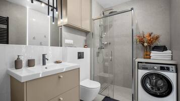 A modern bathroom with a glass shower enclosure, a built-in washing machine, and a vanity with a mirror.
