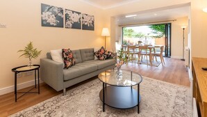 Un salon avec un canapé gris, une table basse en verre et un tapis à motifs. On aperçoit un coin repas avec des chaises en bois et une table, visibles à travers une porte ouverte.