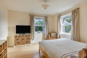 A bedroom with a bed, a TV on a white cabinet, wicker baskets, a chair, and windows with curtains.