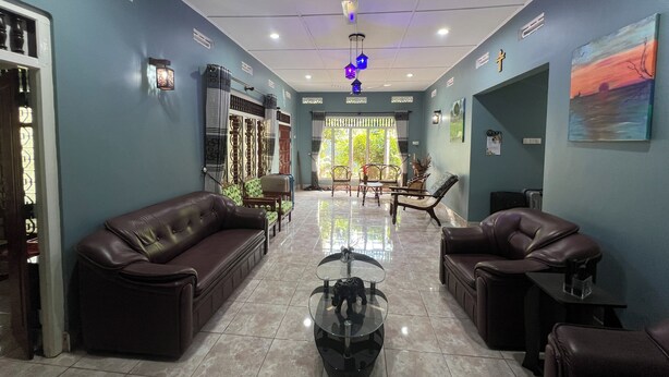 A spacious lobby with a high ceiling, a ceiling fan, and a large window providing natural light. There are leather sofas and a glass coffee table in the center.