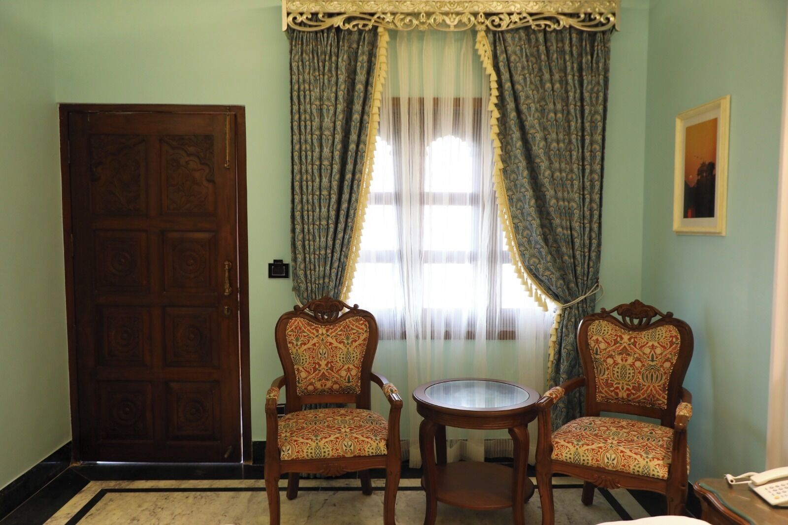 Una habitación con dos sillones decorativos, una mesita redonda pequeña, una puerta de madera y un ventanal con cortinas estampadas.