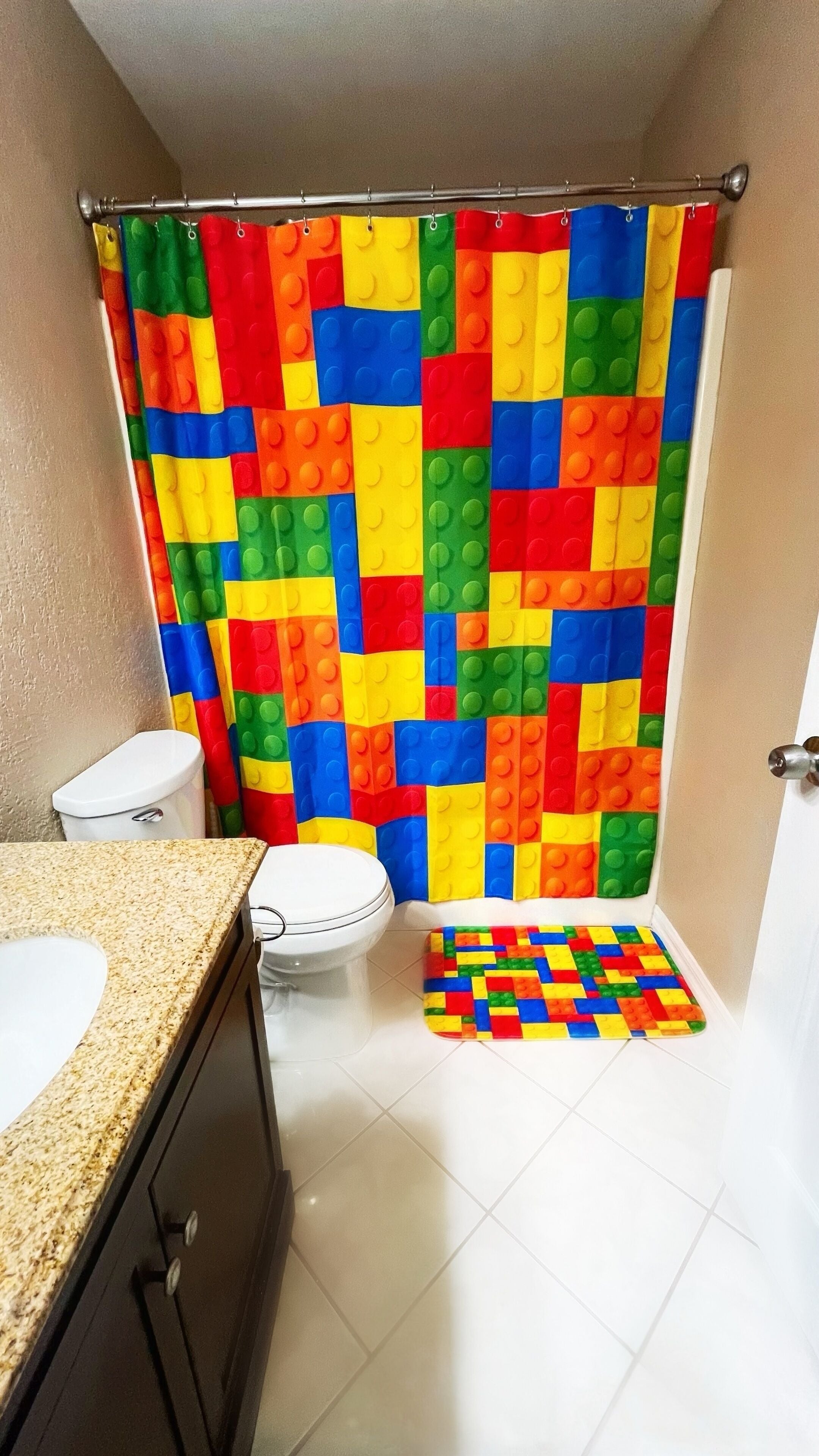 Een badkamer met een douchegordijn in een kleurrijk LEGO-patroon, een wit toilet en een klein vloerkleedje met een LEGO-patroon.