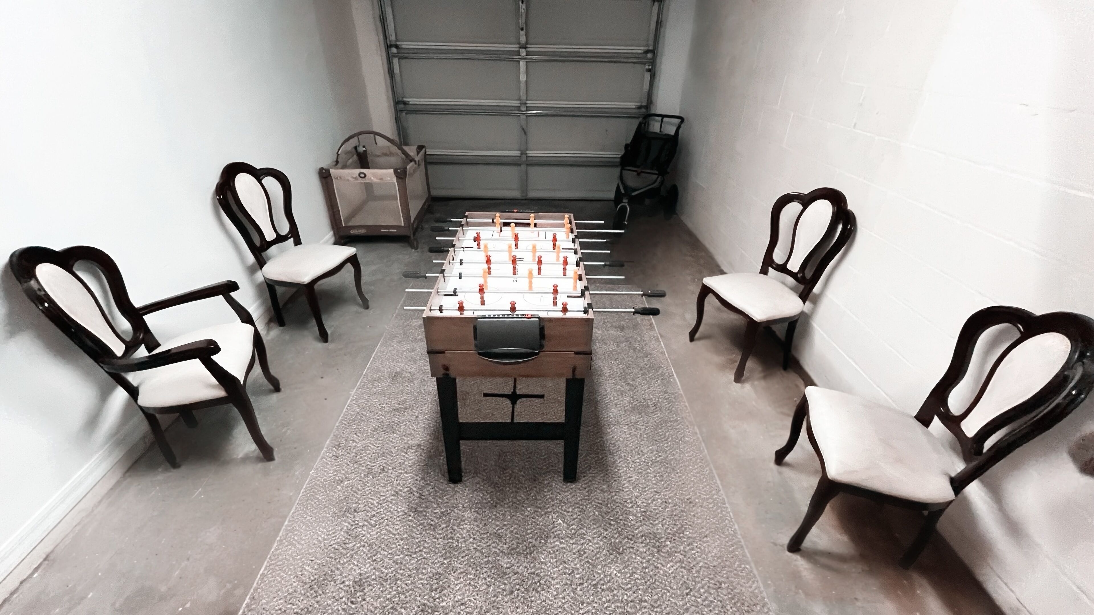 A foosball table surrounded by four chairs in a narrow space with a garage door in the background.