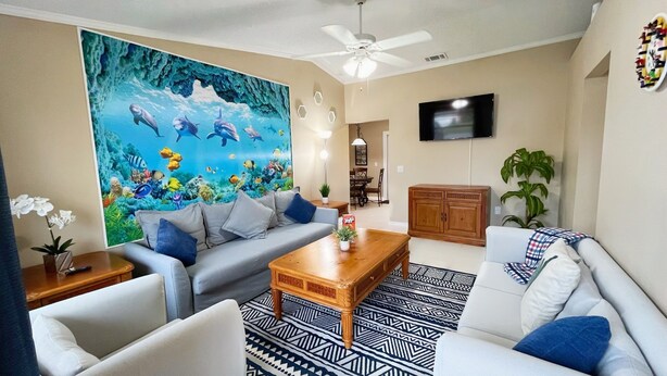 A living room with a large mural of marine life, a ceiling fan, a flat-screen TV, and a dining area in the background. - BlockHaven near Legoland & Disney (Winter Haven)