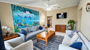 A living room with a large mural of marine life, a ceiling fan, a flat-screen TV, and a dining area in the background. - BlockHaven near Legoland & Disney (Winter Haven)